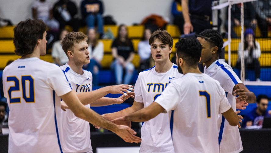 The TMU men's volleyball team in white jerseys huddles after losing a point