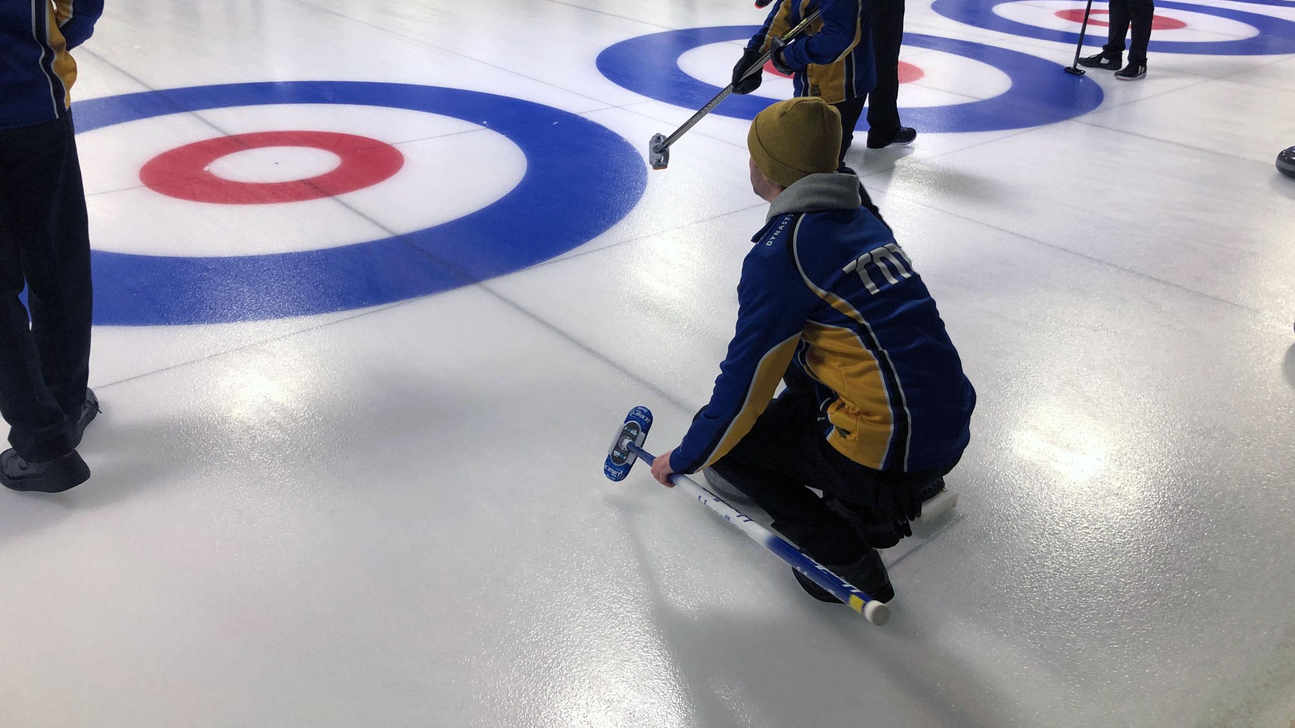A TMU men's curler in a blue and gold jacket prepares for their next shot