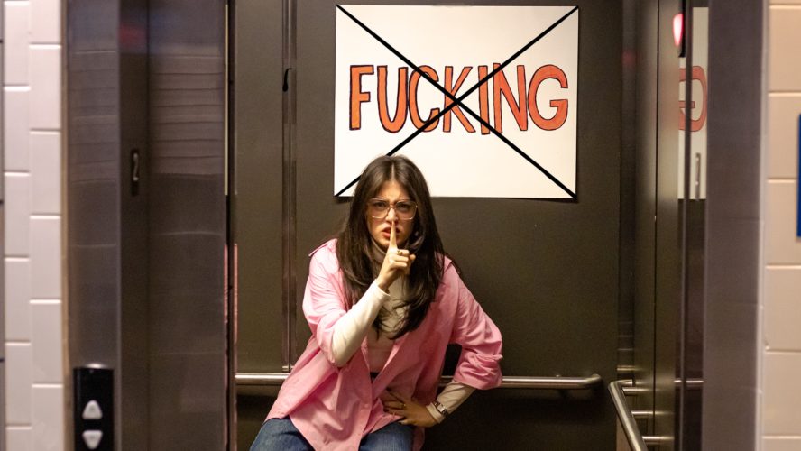 A woman shushing while standing in an elevator. A sign that says "fucking" with an "X" over it hangs above her head.