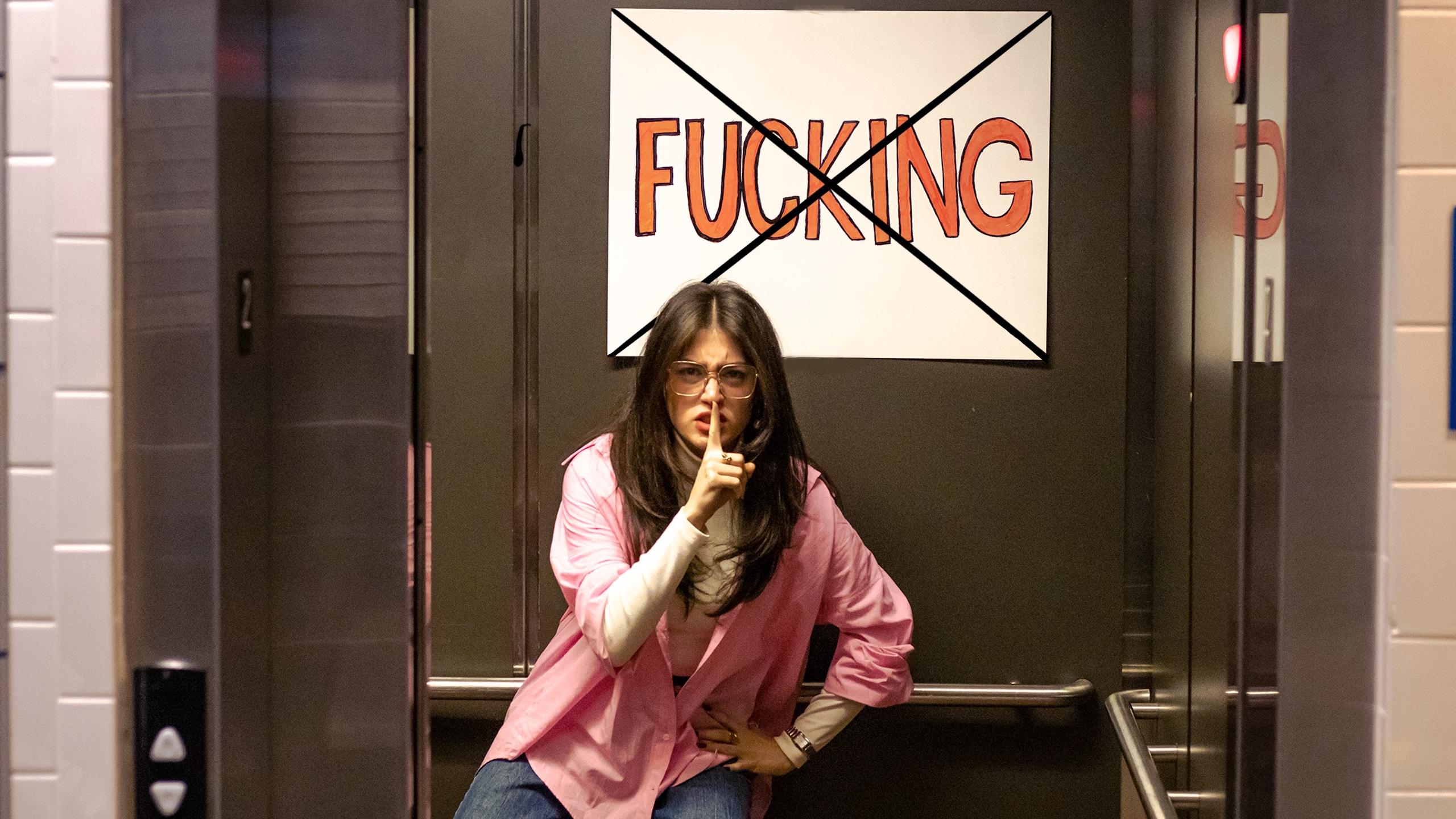 A woman shushing while standing in an elevator. A sign that says "fucking" with an "X" over it hangs above her head.