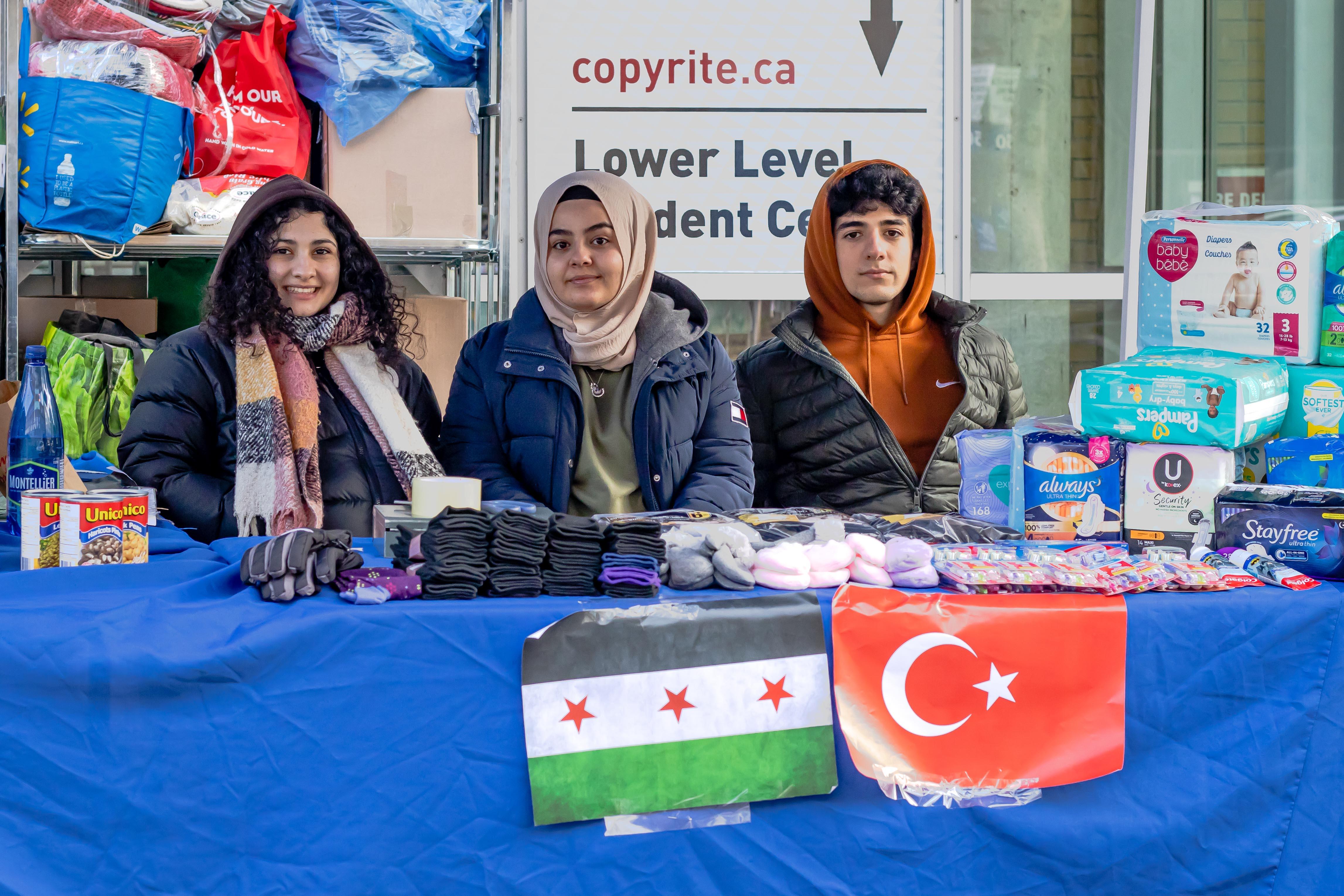 Three people sitting behind a stand, surrounded by canned goods, diapers, socks and other essential household items. A Syria and Turkey flag is taped to the table.