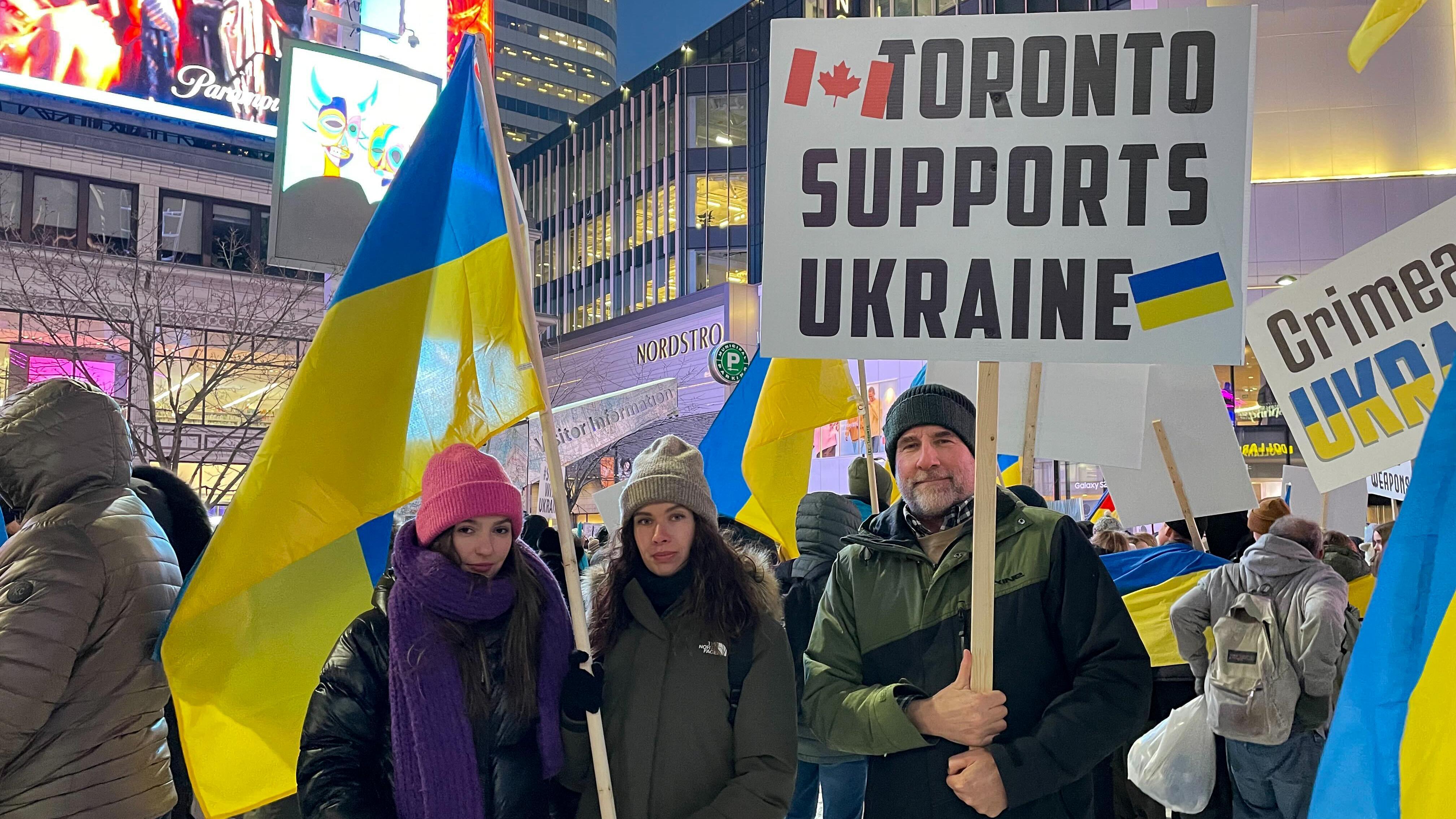 Three people looking at the camera at the rally. One holds a sign stating "stand with Ukraine," and another holds a flag.