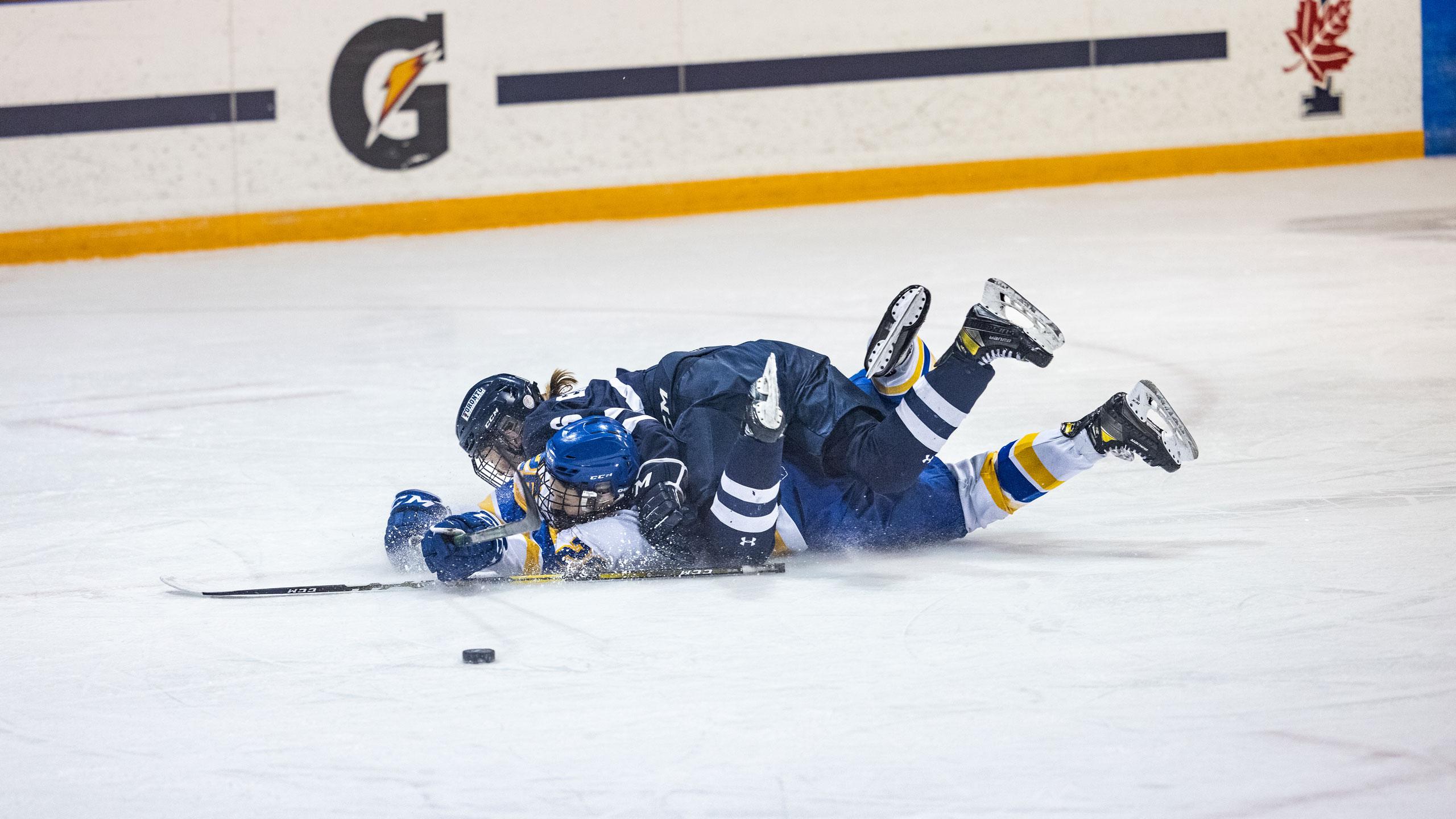 A TMU women's hockey player and a U of T women's hockey player get tangled up in a pile near the puck