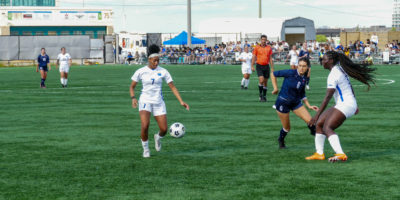 TMU Bold player Taliyah Walker prepares to kick bouncing ball as other players and fans watch on
