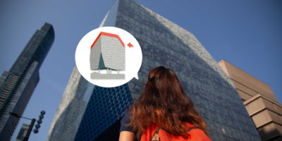 Up-facing angle of a woman facing a tall building wearing an orange backpack