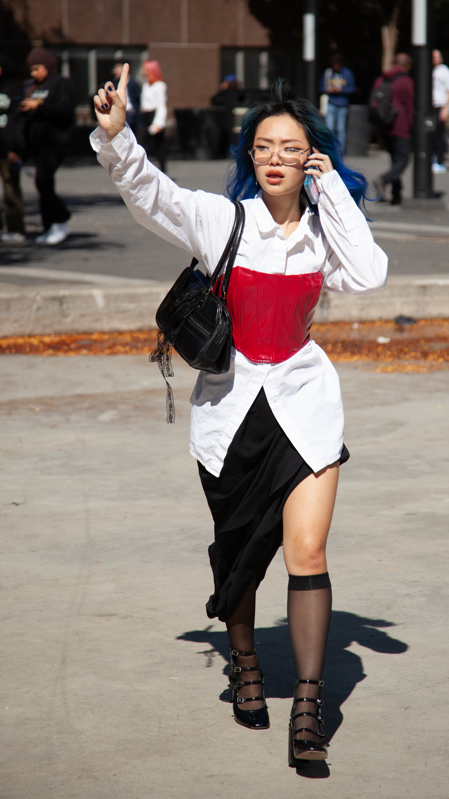 Model walks through campus wearing a bright red corset over button shirt and a sheer dress while pretending to hail a cab