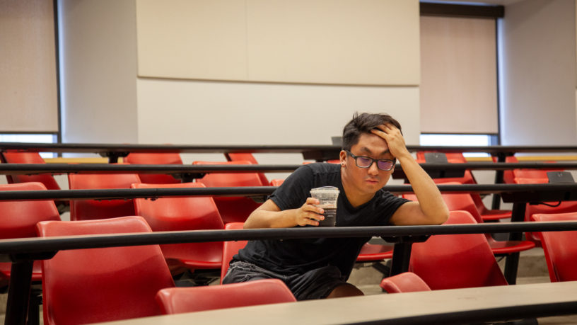 tired-looking student sits alone in a classroom at a desk holding a coffee cup in one hand and holding his forehead in the other hand propped up on the desk