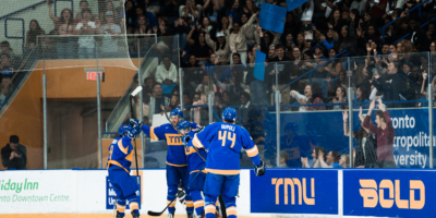 Three TMU Bold men's hockey players celebrate with the sold-out crowd in the background