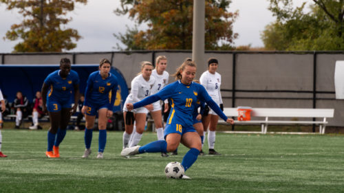 TMU Bold women's soccer player begins to take penalty kick with her teammates and Laurentian Voyageurs players watching from behind her