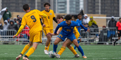 TMU men's soccer player Bilal Reslan battles for the soccer ball with multiple Laurentian Voyageurs players as teammates and opponents watch from behind