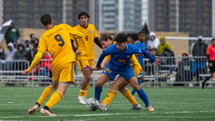 TMU men's soccer player Bilal Reslan battles for the soccer ball with multiple Laurentian Voyageurs players as teammates and opponents watch from behind