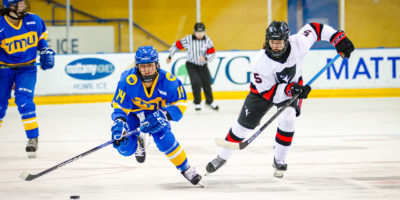TMU Bold women's hockey player Megan Bergmanis and a Carleton Ravens player chase after a loose puck
