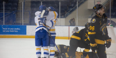 Two players of the TMU men's hockey team hug in celebration of a goal in front of the Waterloo Warriors goaltender