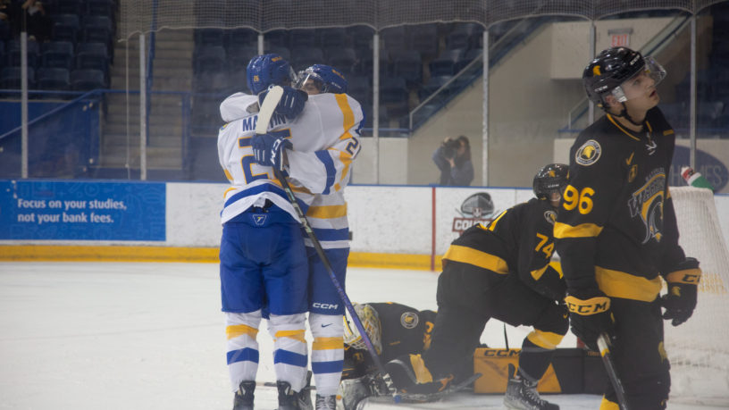 Two players of the TMU men's hockey team hug in celebration of a goal in front of the Waterloo Warriors goaltender