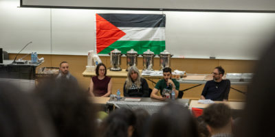 Attendees watch on as the speakers of the Panel for Palestine event speak. In the background a Palestinian flag hangs on a whiteboard.