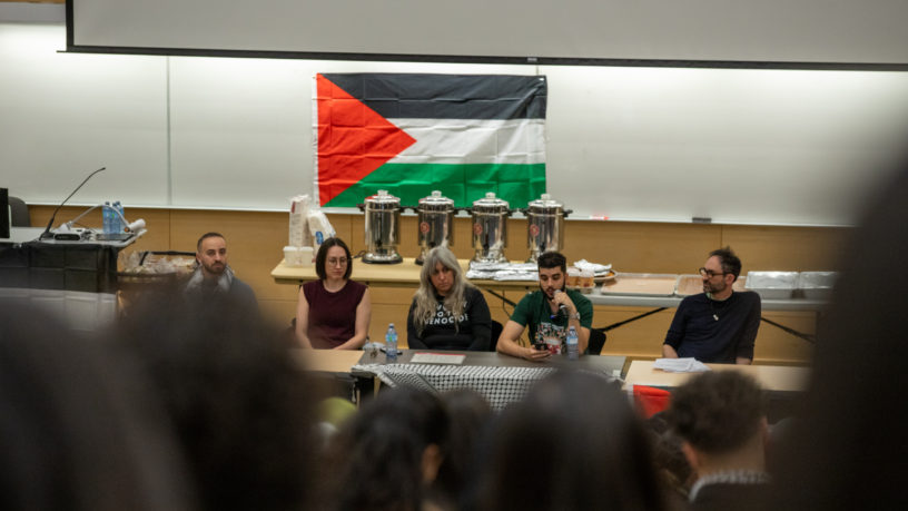 Attendees watch on as the speakers of the Panel for Palestine event speak. In the background a Palestinian flag hangs on a whiteboard.