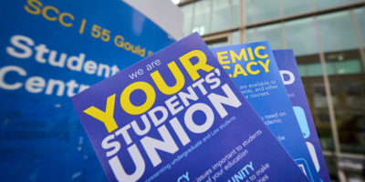 Flyers in blue, yellow and white being held up in front of SCC building sign