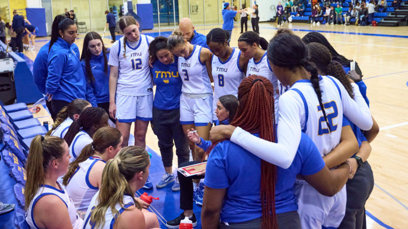 The TMU women's basketball team huddles around head coach Carly Clarke during a timeout