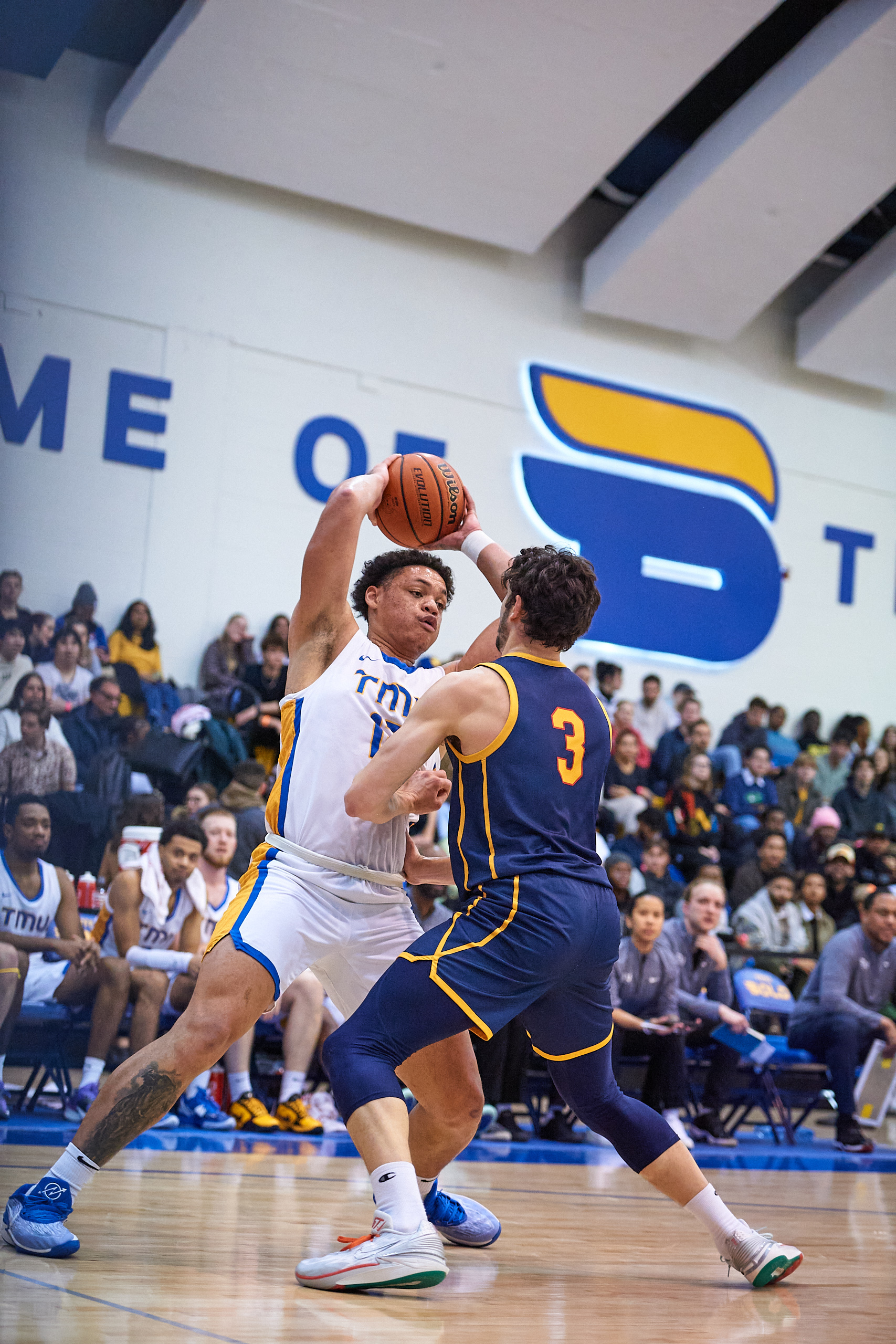 TMU men's basketball player Aaron Rhooms holds the ball over his head while being guarded by Queen's player