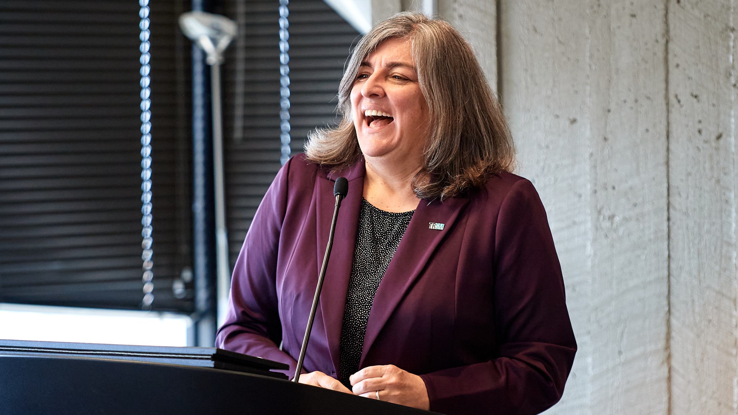 Woman laughing while standing at podium, wearing purple blazer