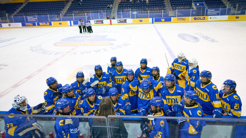 Members of the TMU Bold women's hockey team huddle by the bench