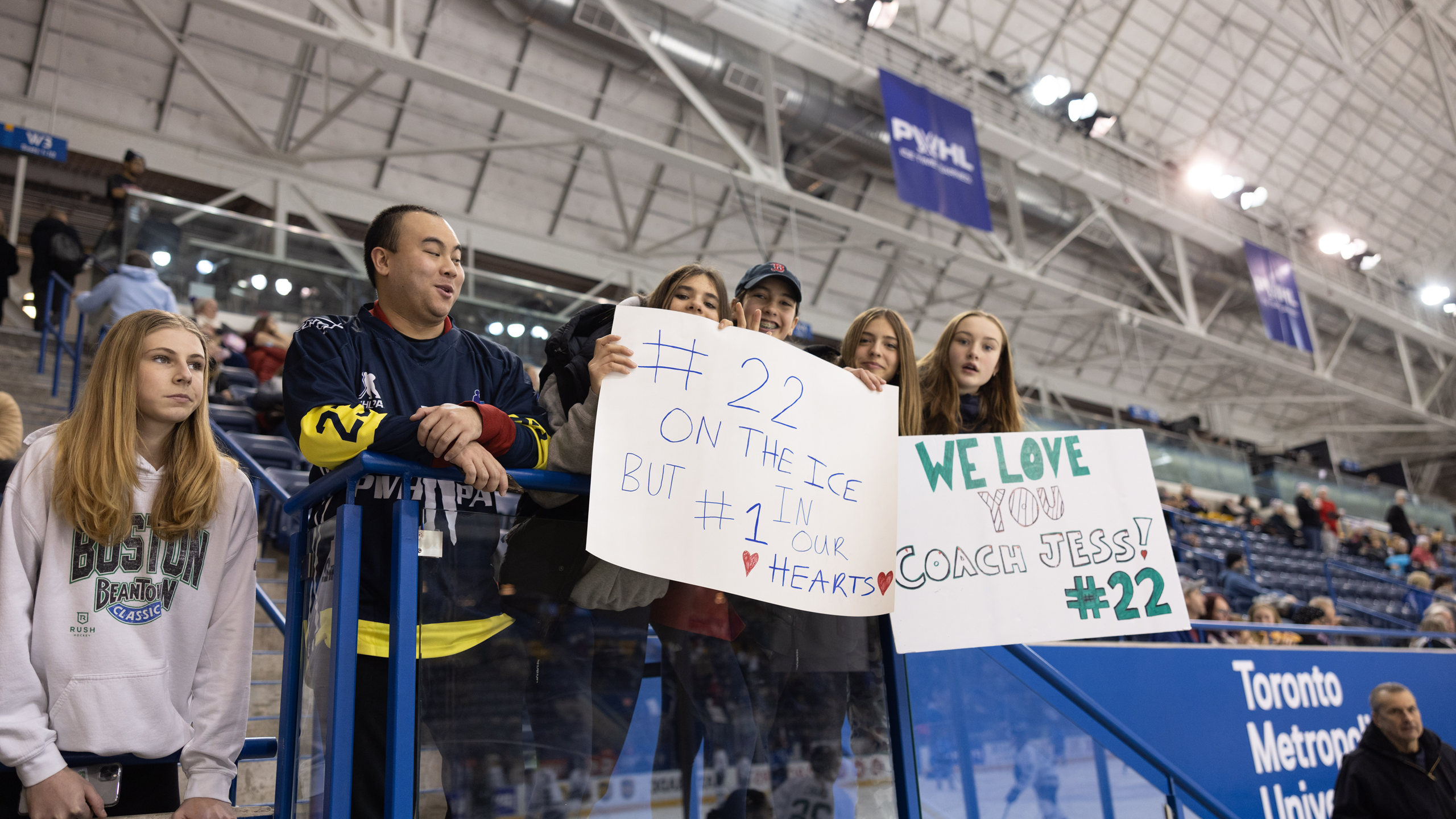 Fans hold up two signs. The first says #22 on the ice but #1 in our hearts. The second sign says "We love you coach Jess #22"