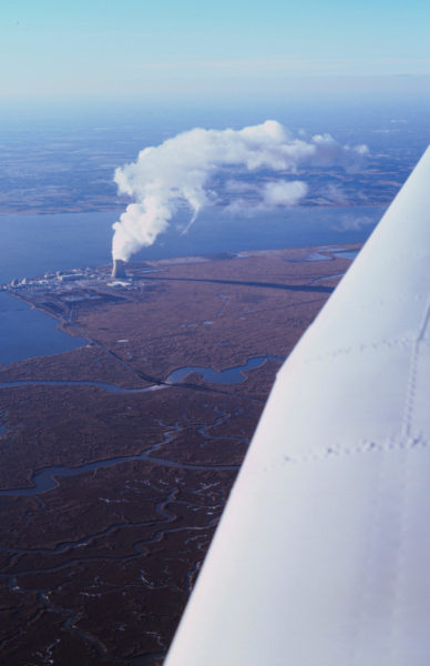 The wing of a plane in the air is on the right side with the background of fields and a plant with smoke.