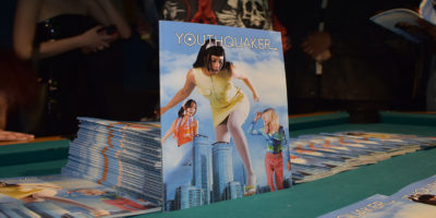 A stack of Youthquaker magazines lays atop a billiards table with one of the magazines lays propped against the stack, showcasing its cover of a woman towering over skyscrapers.