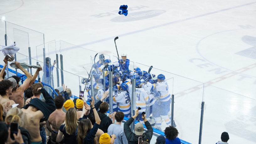 The TMU Bold men's hockey team celebrates their win in a huddle as the crowd cheers them on