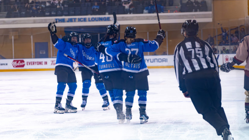 Two PWHL Toronto teammates hug each other as two other skate towards them in celebration of a goal