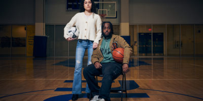 A man sits on a chair in the middle of a basketball court with a woman standing next to him