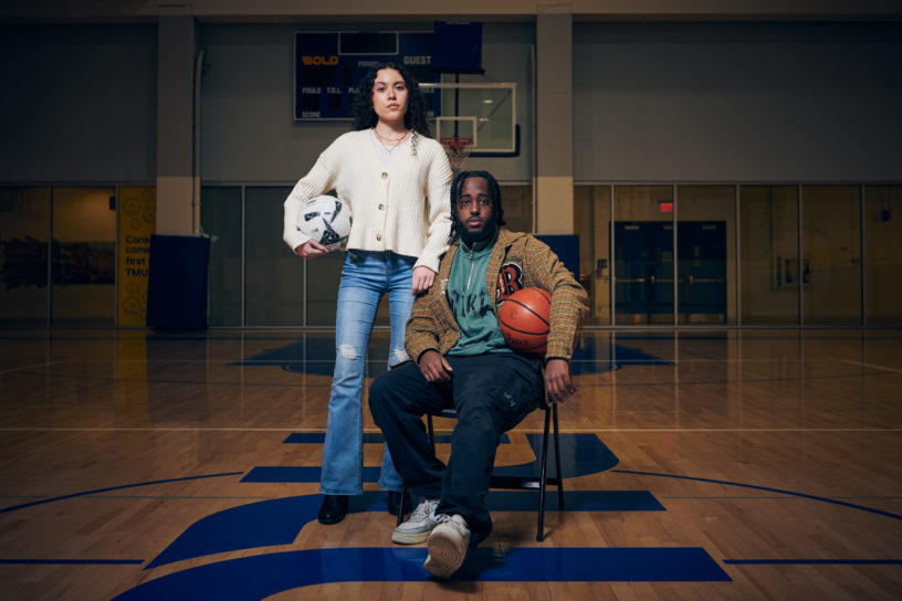 A man sits on a chair in the middle of a basketball court with a woman standing next to him