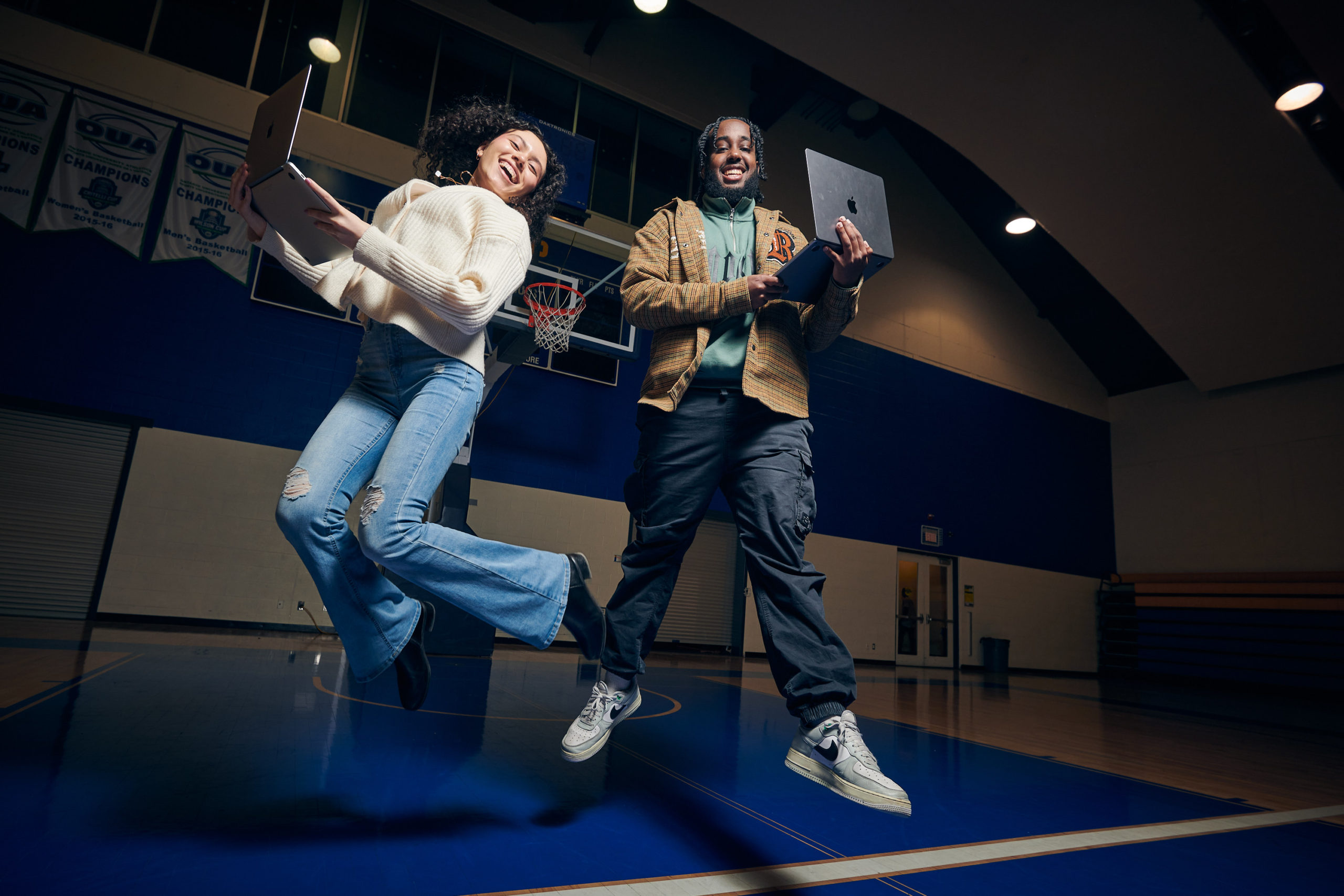 Two people jump holding laptops on a basketball court