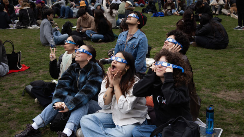 Five people sitting on grass look up to sky with solar eclipse glasses on