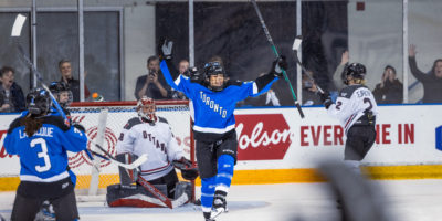 PWHL Toronto's Sarah Nurse celebrates a goal with her stick in the air. Fans react with glee in the background