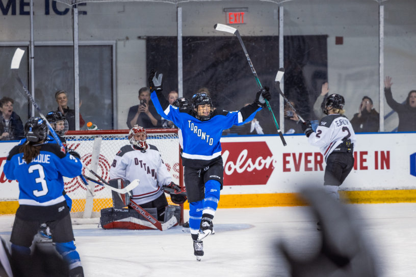PWHL Toronto's Sarah Nurse celebrates a goal with her stick in the air. Fans react with glee in the background