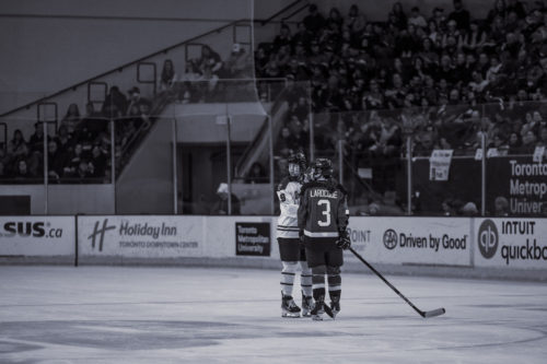 A black and white photo shows a player in a PWHL Toronto uniform and a player in a PWHL Ottawa uniform speaking at centre ice