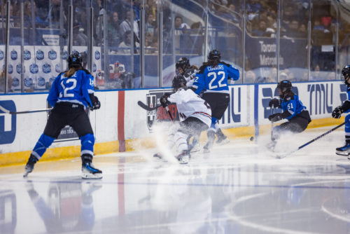 A player in a white PWHL Ottawa uniform stops and sprays snow near the boards and two PWHL Toronto players