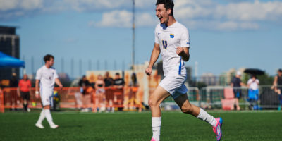 TMU Bold men's soccer player Chris Campoli celebrates a goal with a running fist pump. He is wearing a white jersey