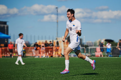 TMU Bold men's soccer player Chris Campoli celebrates a goal with a running fist pump. He is wearing a white jersey