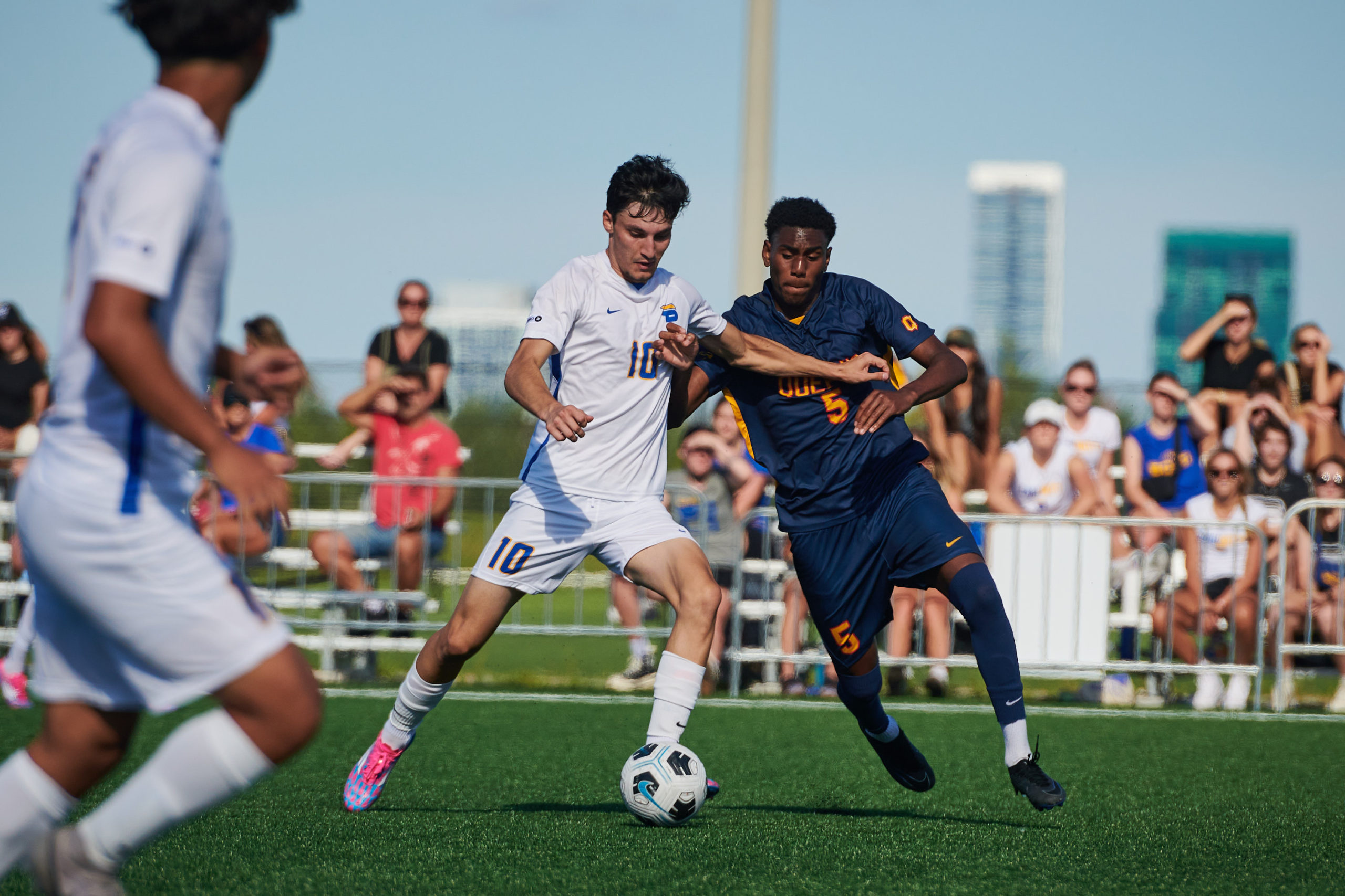 A TMU Bold men's soccer player and a Queen's Gaels men's soccer player battle for a ball