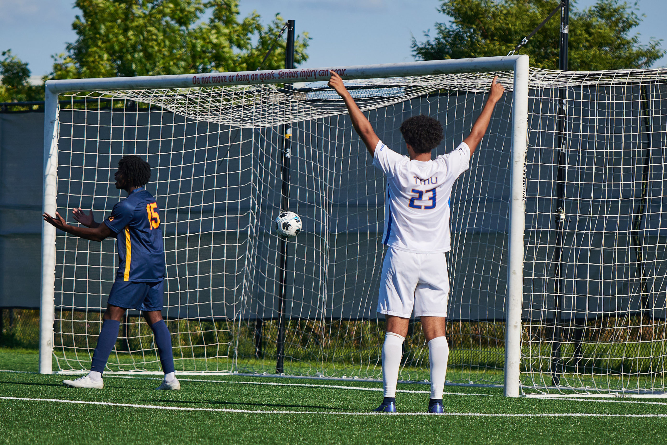 a TMU Bold player in a number 23 jersey celebrates a goal. The ball can be seen going into he net, while a Queen's player lifts his head to the sky