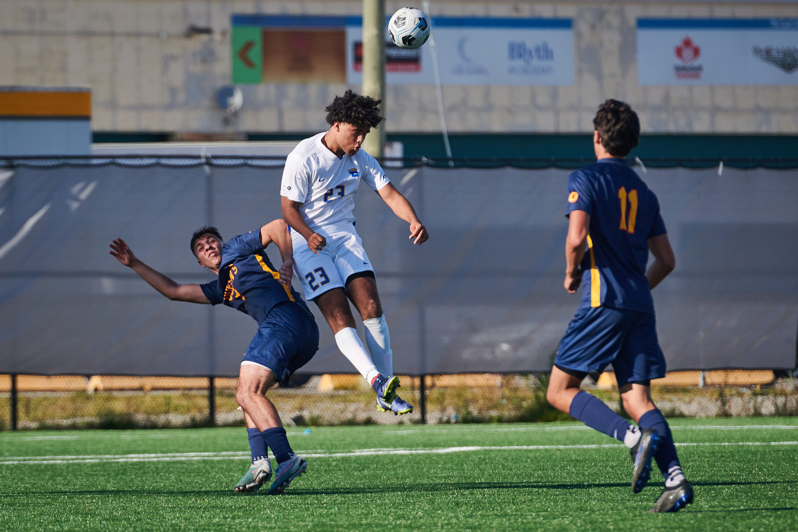 A TMU Bold men's soccer player hits a ball with his head while jumping next to a Queen's Gaels player. Another Queen's player looms