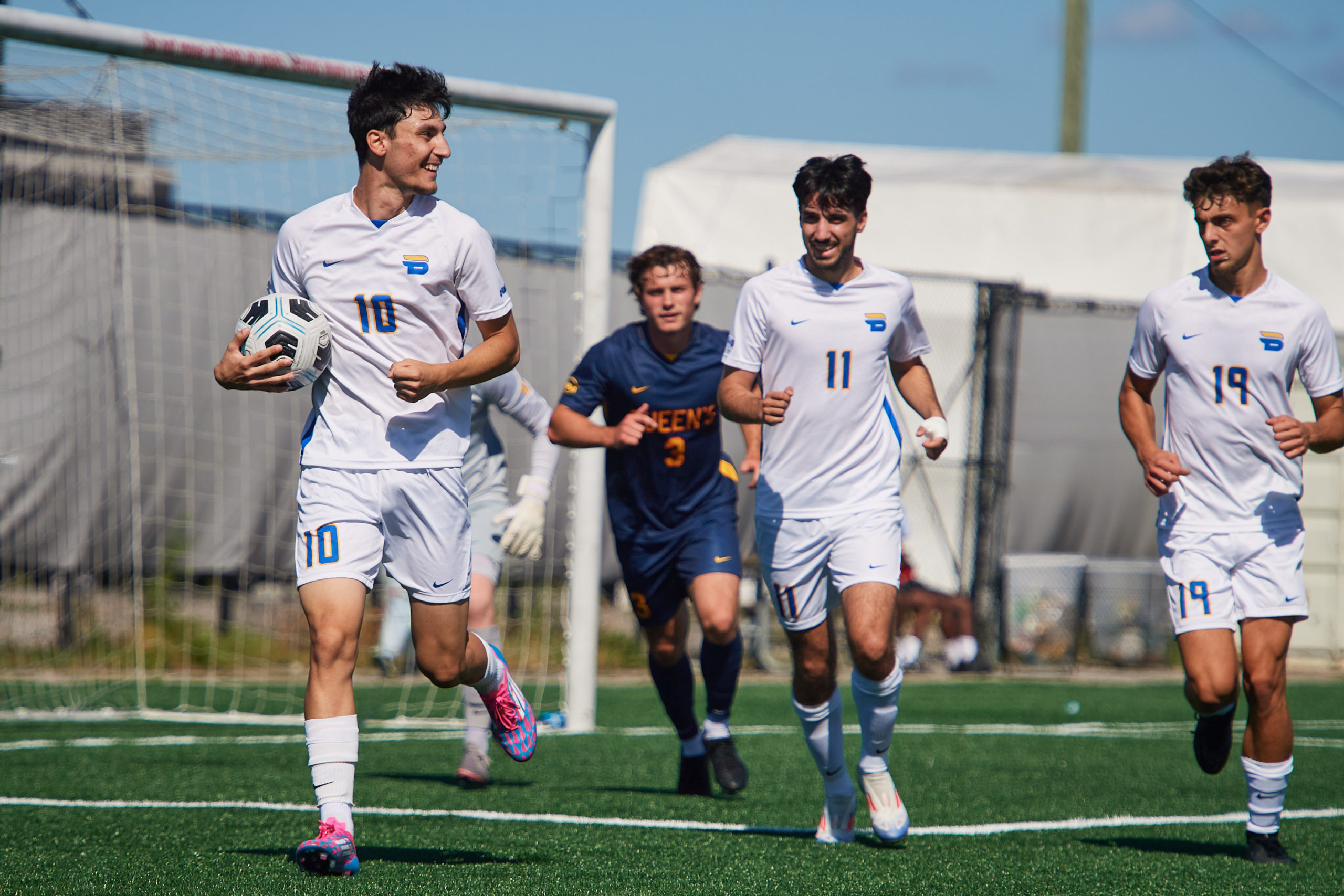 TMU Bold socer player Chris Campoli runs with the ball under his arm after scoring a goal. TMU players follow with a smile, while a Queen's player chases looking less amused