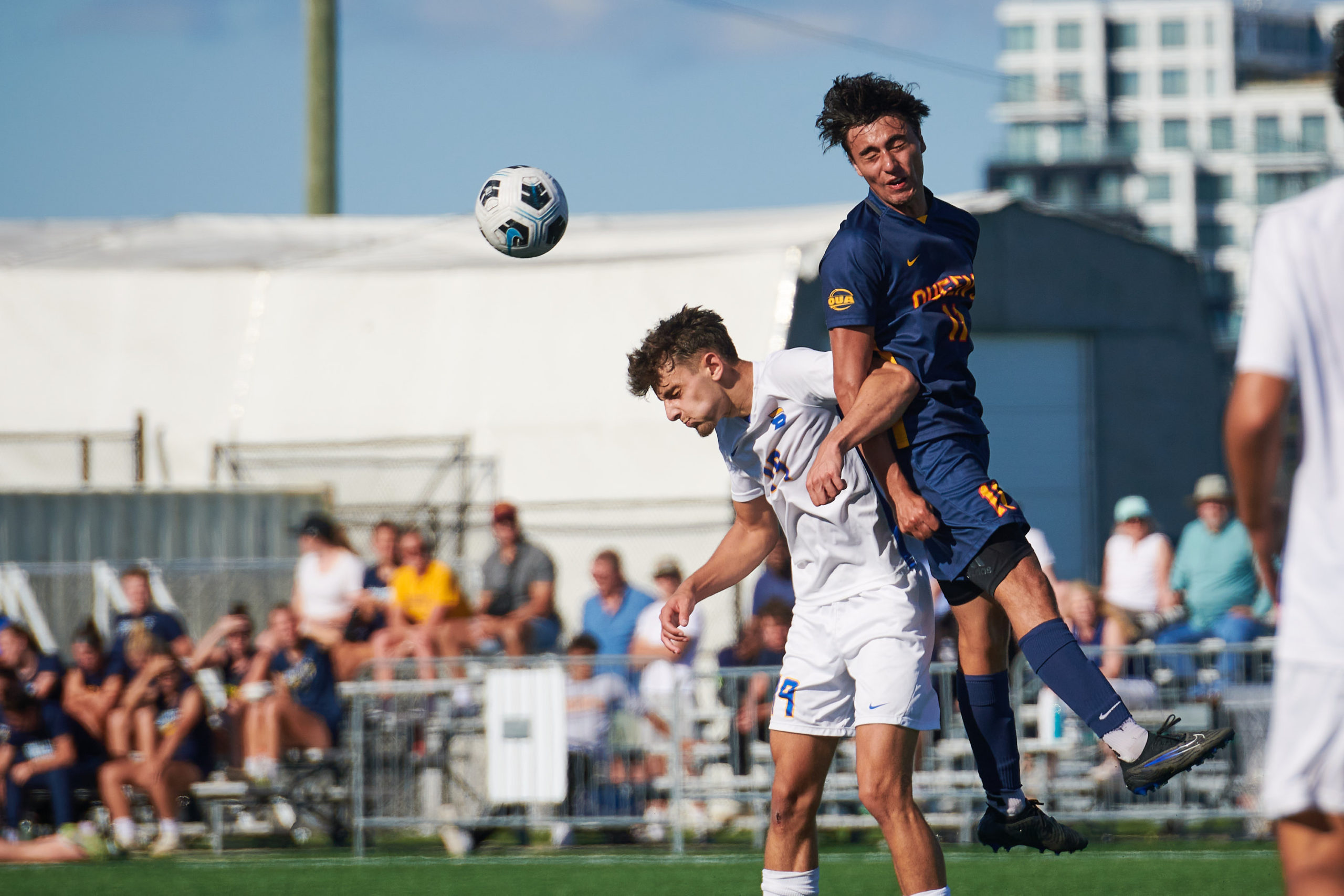 A TMU Bold soccer player hits the ball with his head while a Queen's player jumps over his back