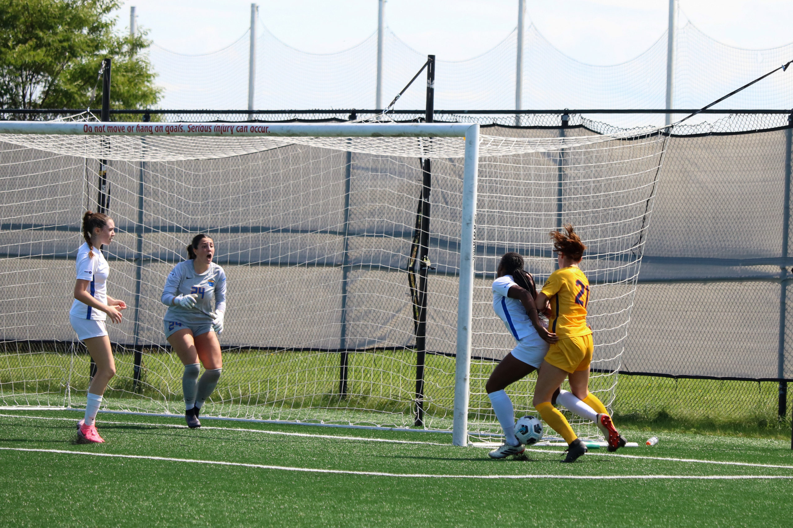 A TMU player and a Queen's player battle for a soccer ball beside the net while two other TMU players watch on. 