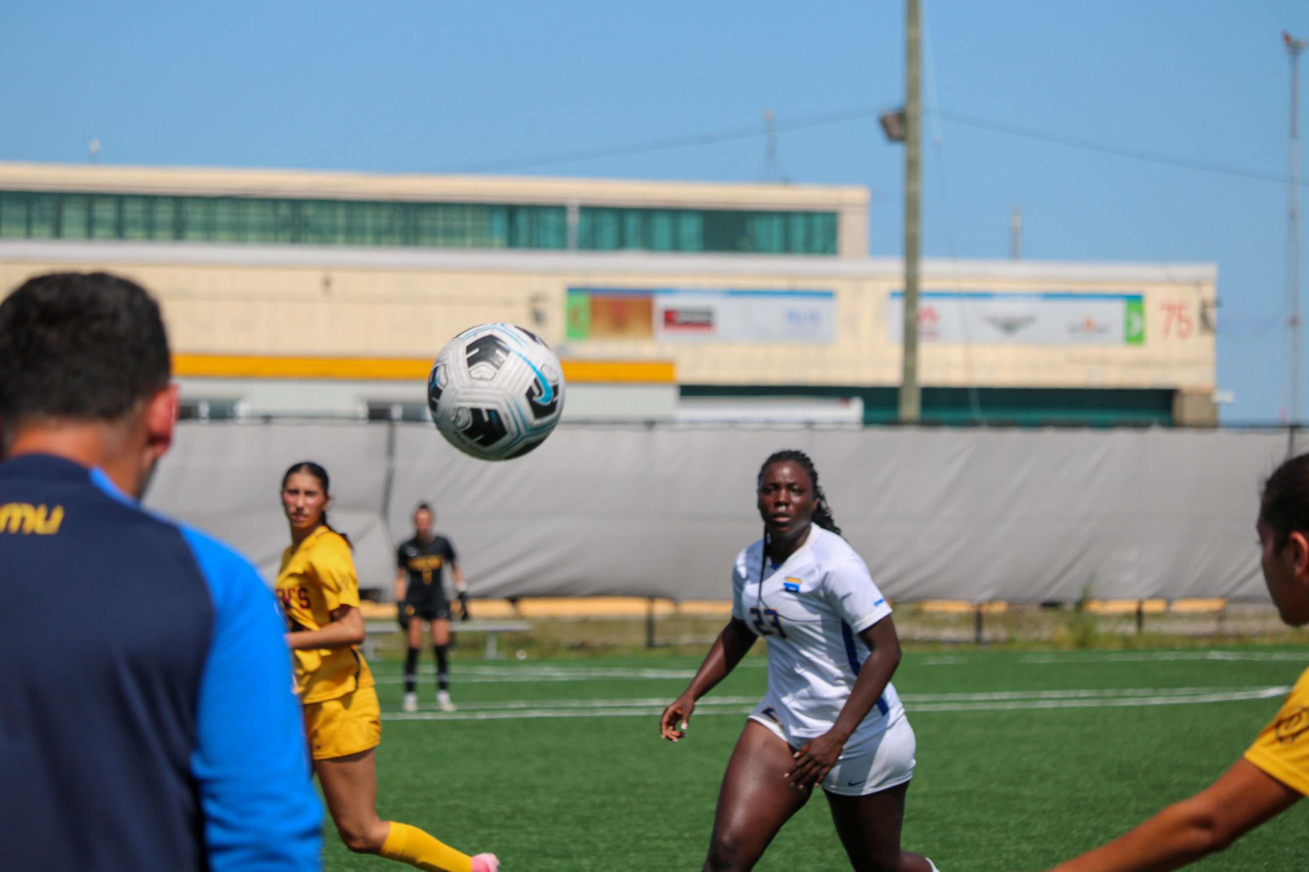 TMU player Aalayah Lully and two Queen's players watch as a soccer ball flies out of bounds near a coach at the sideline.