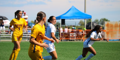 Two TMU Bold women's soccer players in white jerseys chase down a ball alongside two Queen's Gaels players in yellow uniforms