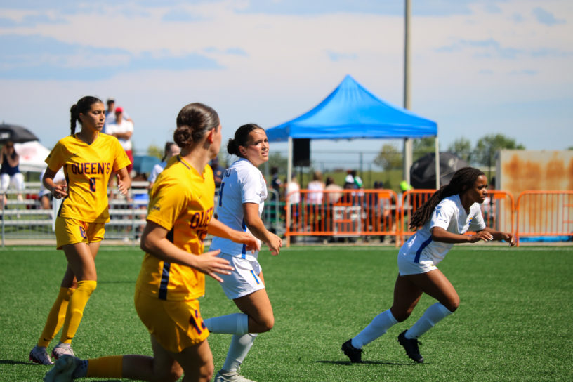 Two TMU Bold women's soccer players in white jerseys chase down a ball alongside two Queen's Gaels players in yellow uniforms