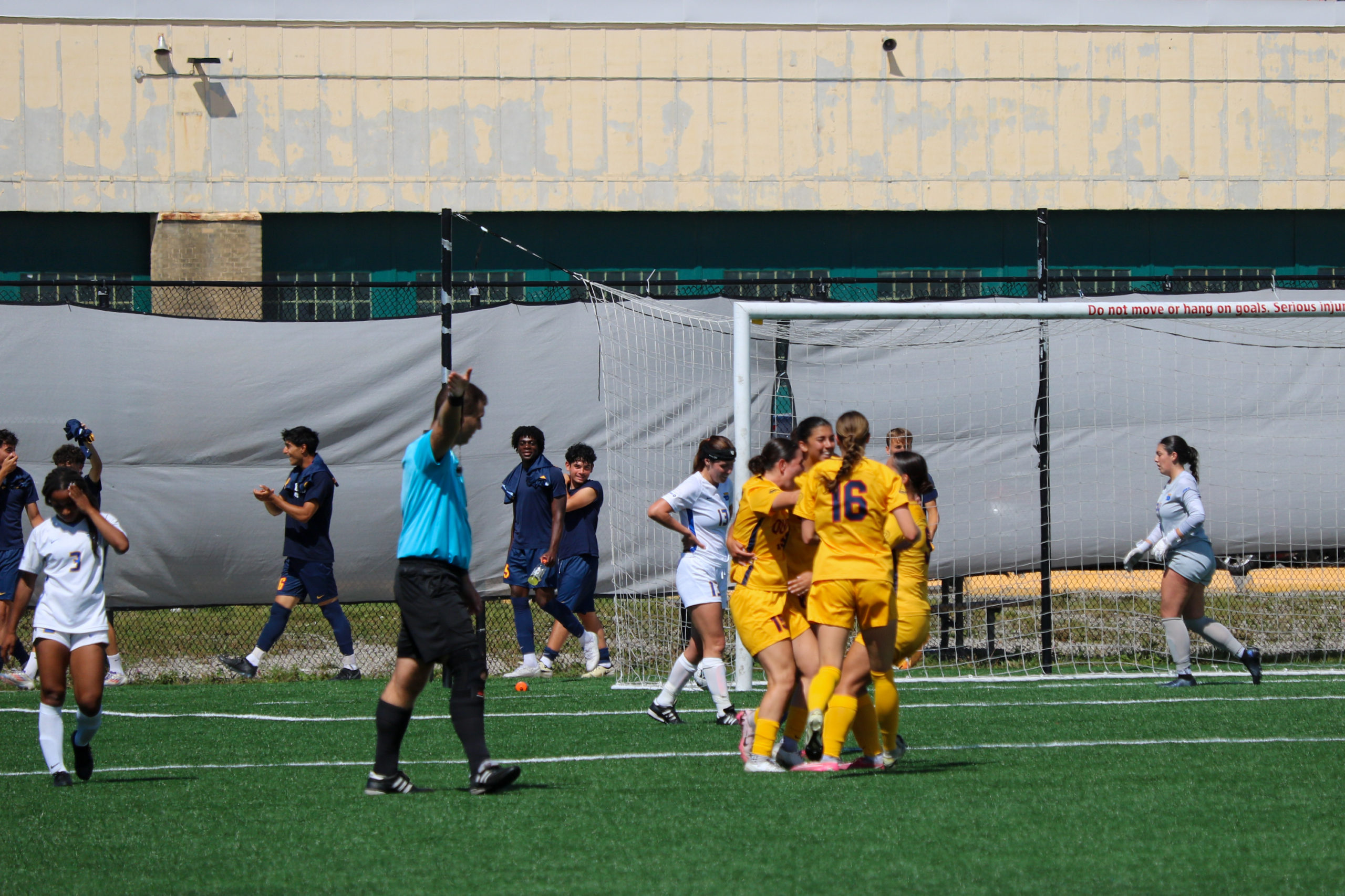 Players for the Queen's Gaels women's soccer team celebrate a goal while TMU Bold players look sullen. A referee has his arm up.